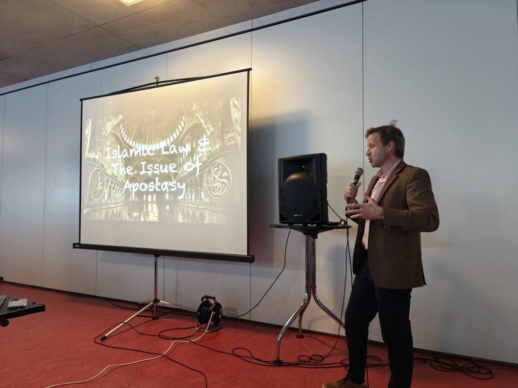 Referent mit Mikrofon neben der Leinwand bei einem Vortrag von Tugend.Club